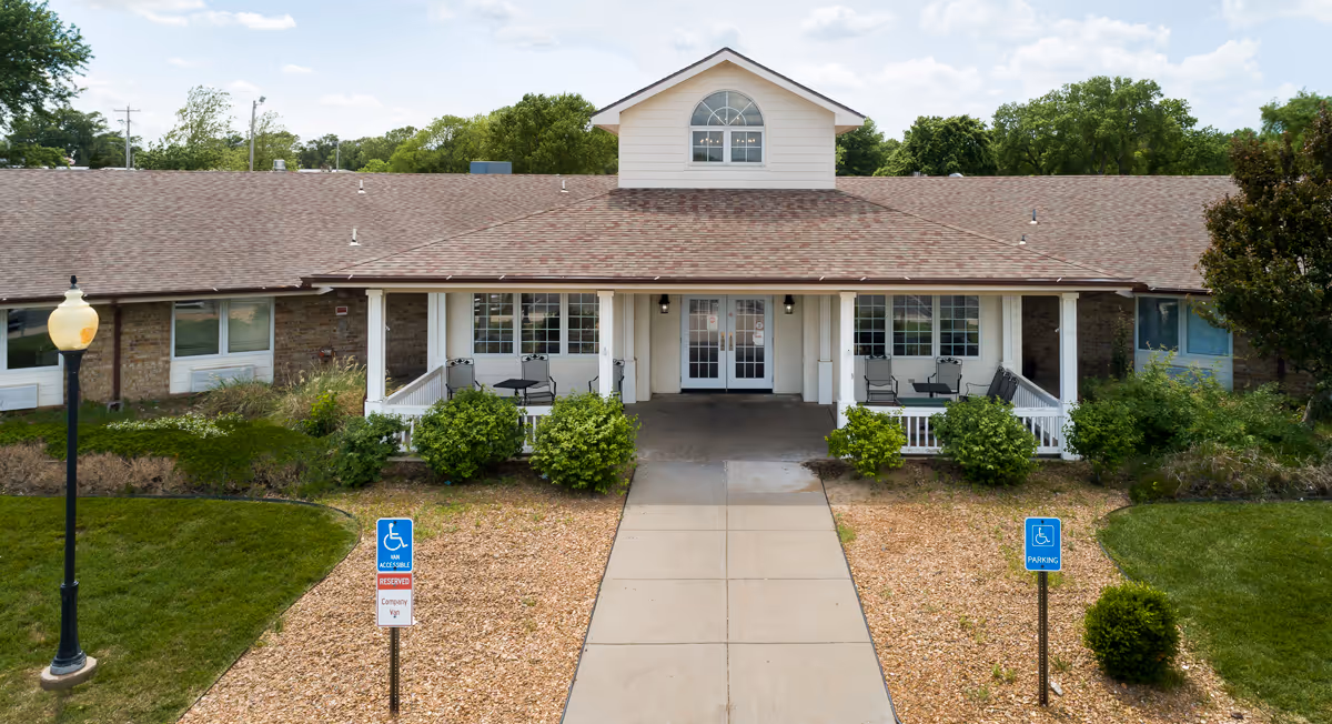 Front exterior view of a single-story building with a covered porch featuring seating areas. There are two handicap parking signs in front of the building, a sidewalk leading to the entrance, and greenery surrounding the area under a partly cloudy sky.