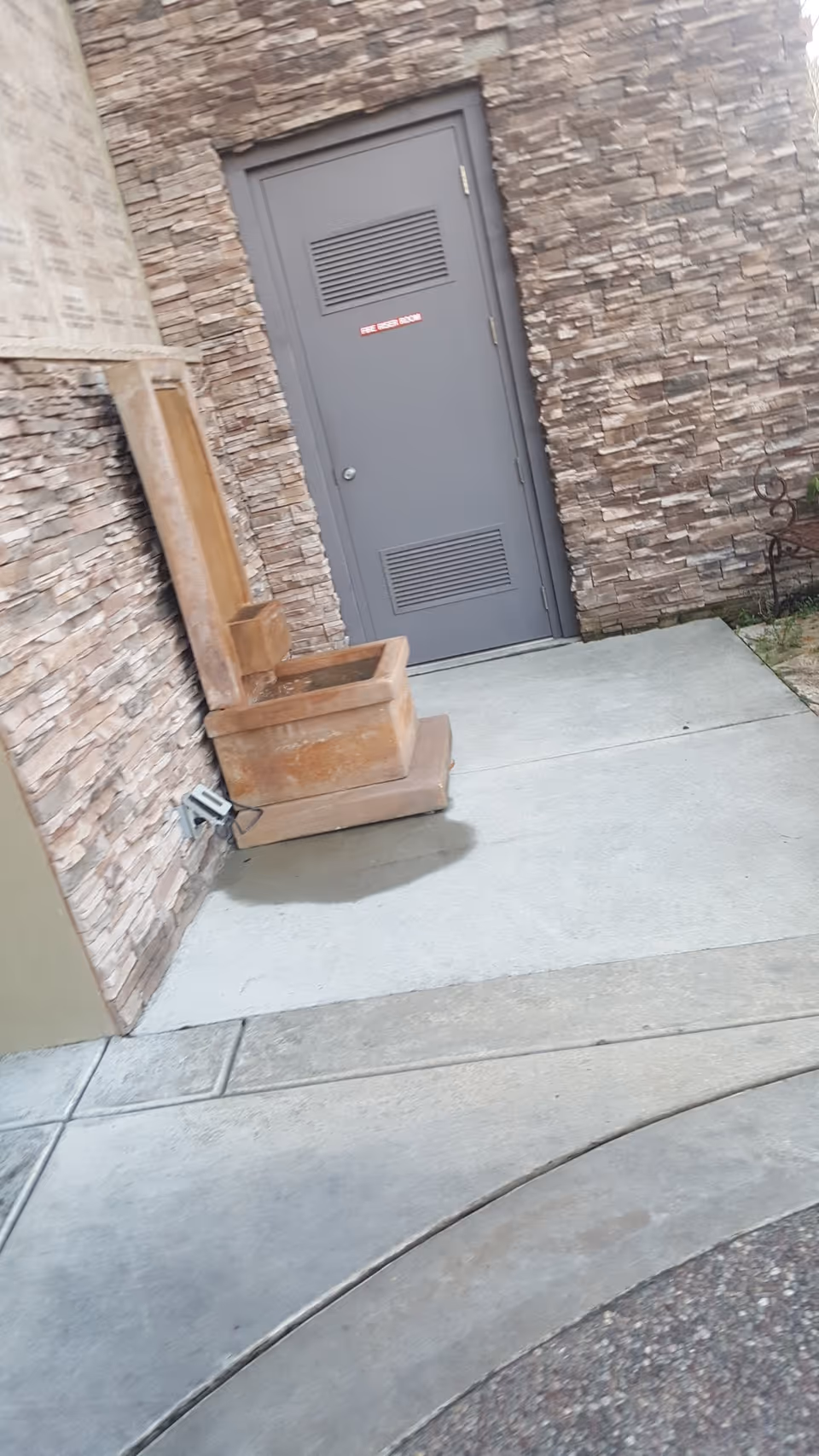 Exterior view of a stone wall entrance with a gray door and concrete walkway.