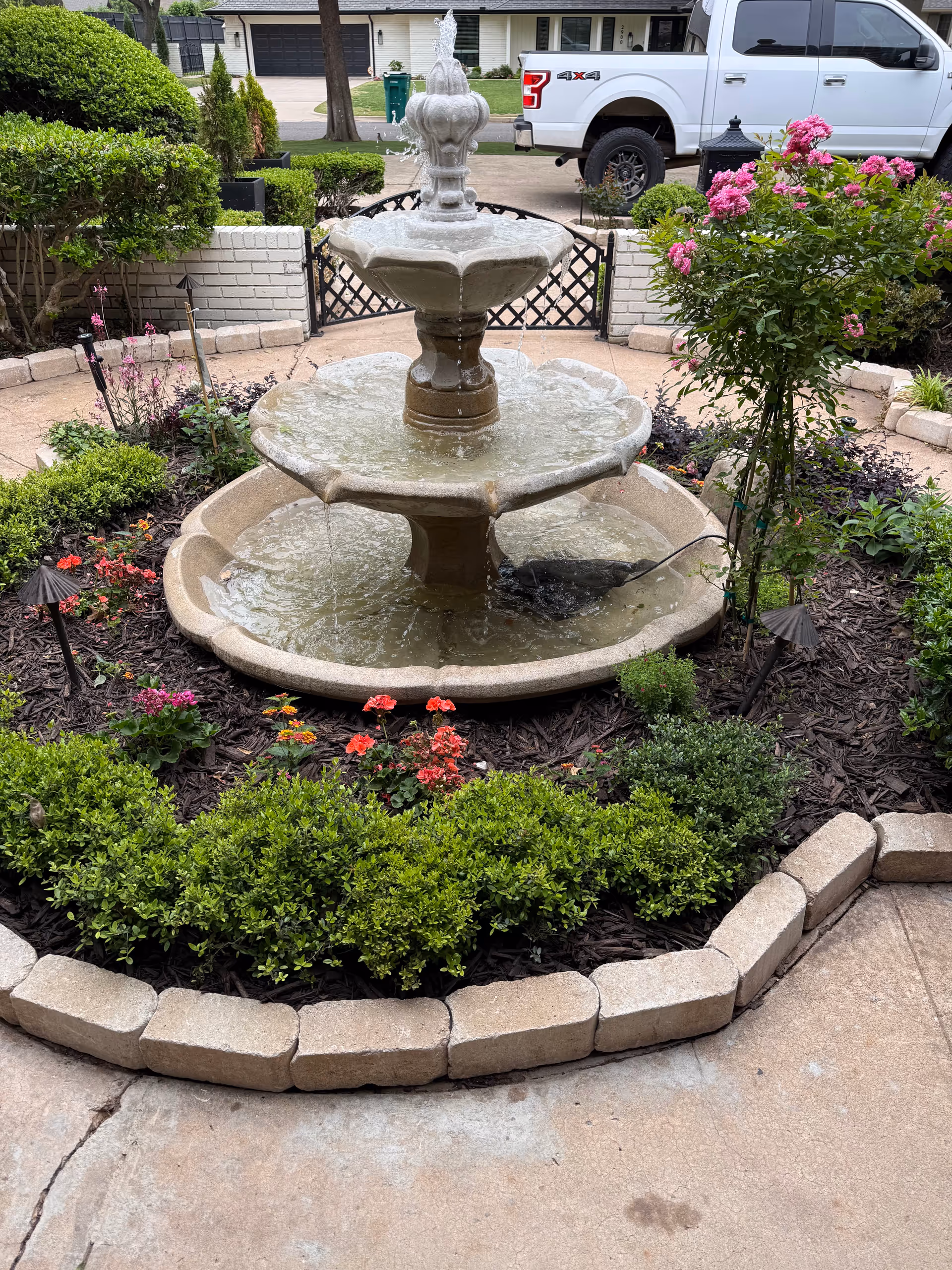 Stone tiered fountain in a landscaped courtyard with blooming flowers and trimmed hedges.