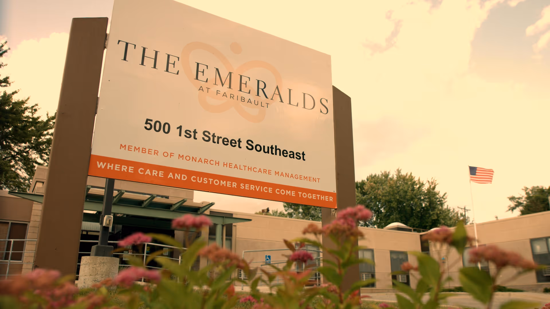 Outdoor view of a sign for The Emeralds at Faribault, located at 500 1st Street Southeast, with a building and American flag in the background and flowers in the foreground.