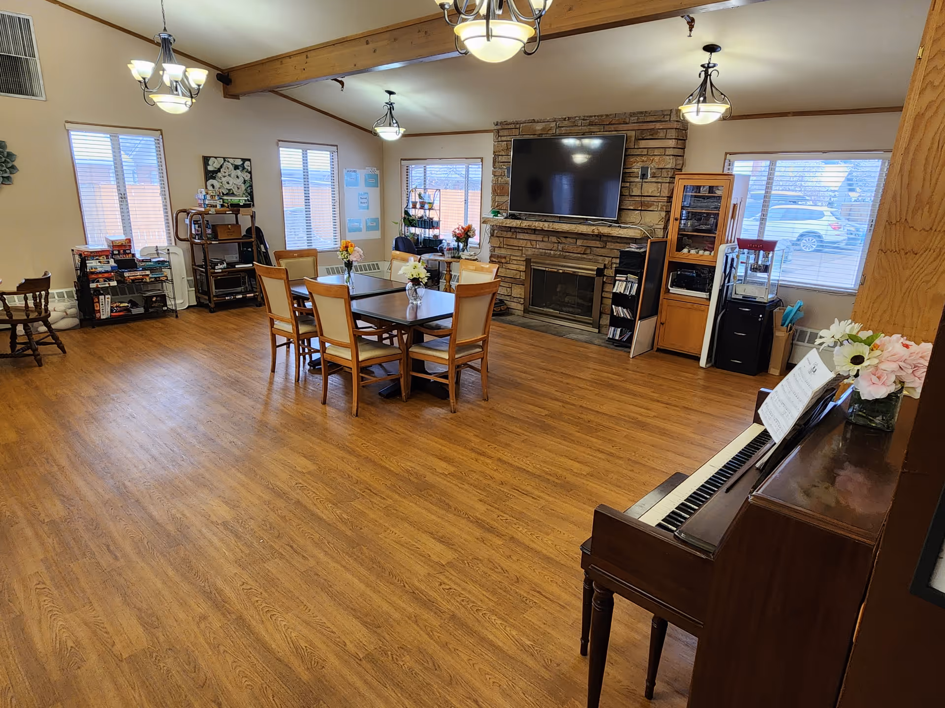 Bright senior living common area with dining table, piano, fireplace, TV, and large windows.