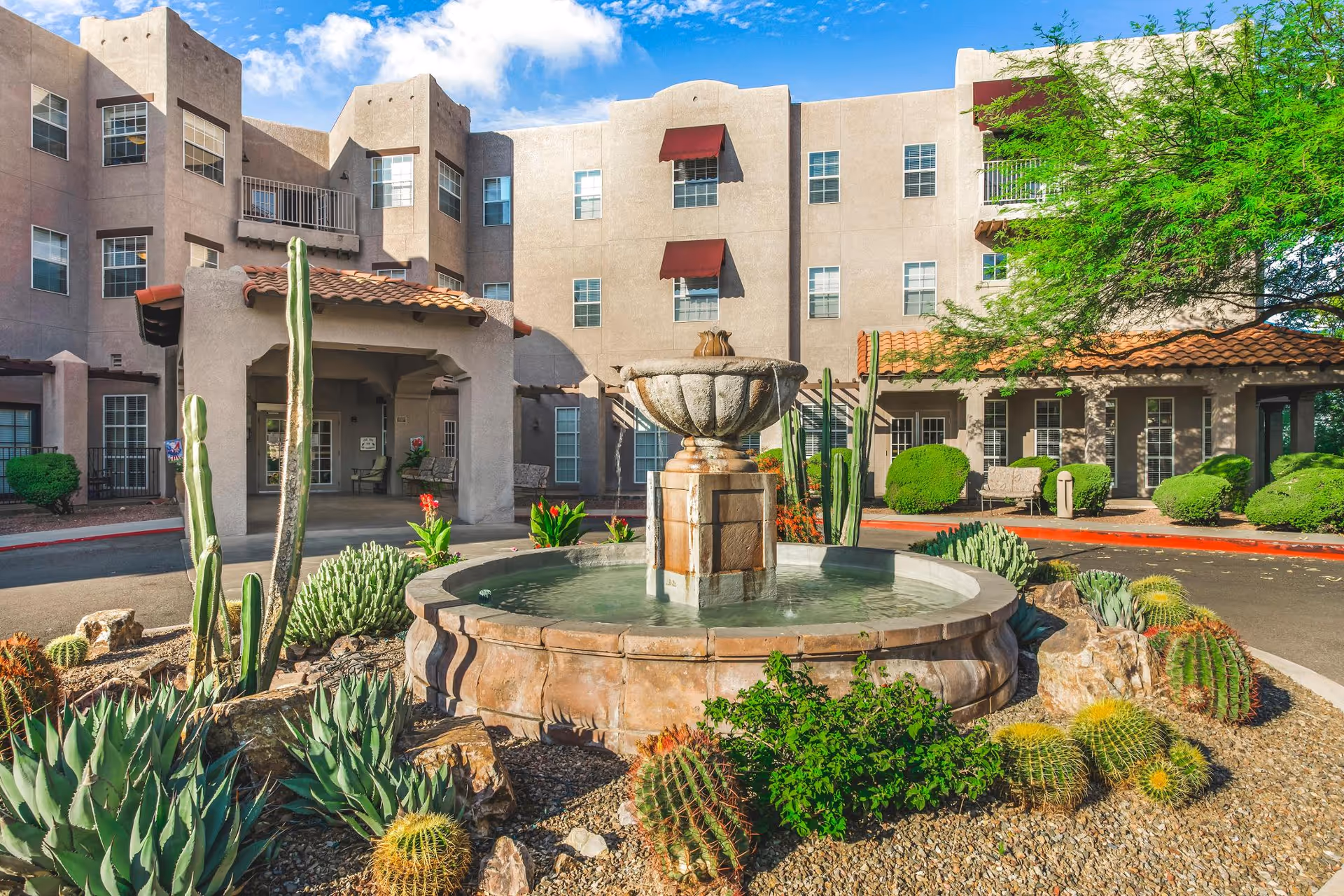 Exterior view of The Fountains at La Cholla senior living facility showing a circular stone water fountain surrounded by desert plants and cacti in the foreground, with a three-story beige building and a covered entrance in the background under a blue sky with some clouds.