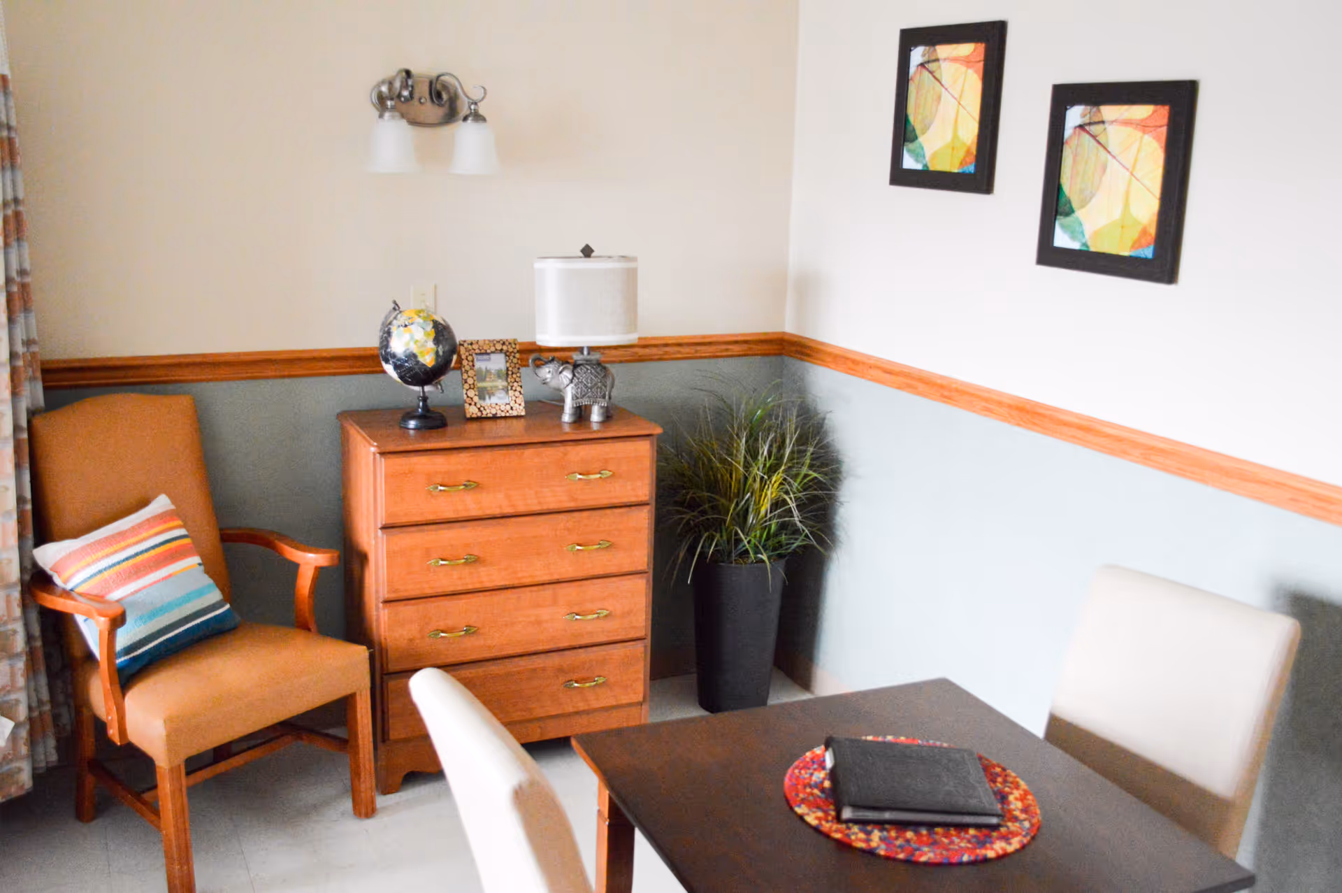 A cozy corner of a room with a wooden chest of drawers topped with a globe, a framed photo, and a lamp shaped like an elephant. Next to the chest is a potted plant. There is an upholstered armchair with a striped pillow on the left and a dark wooden table with two beige chairs on the right. Two colorful abstract paintings hang on the wall above the chest of drawers.