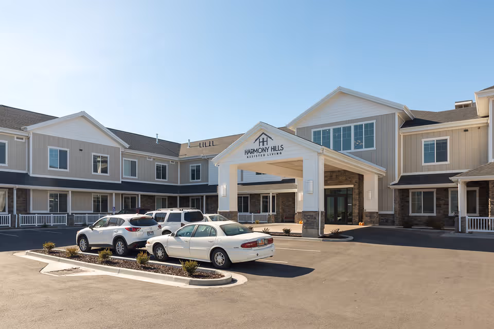 Entrance and parking area of a two-story assisted living building with a covered drop-off portico and several parked cars.