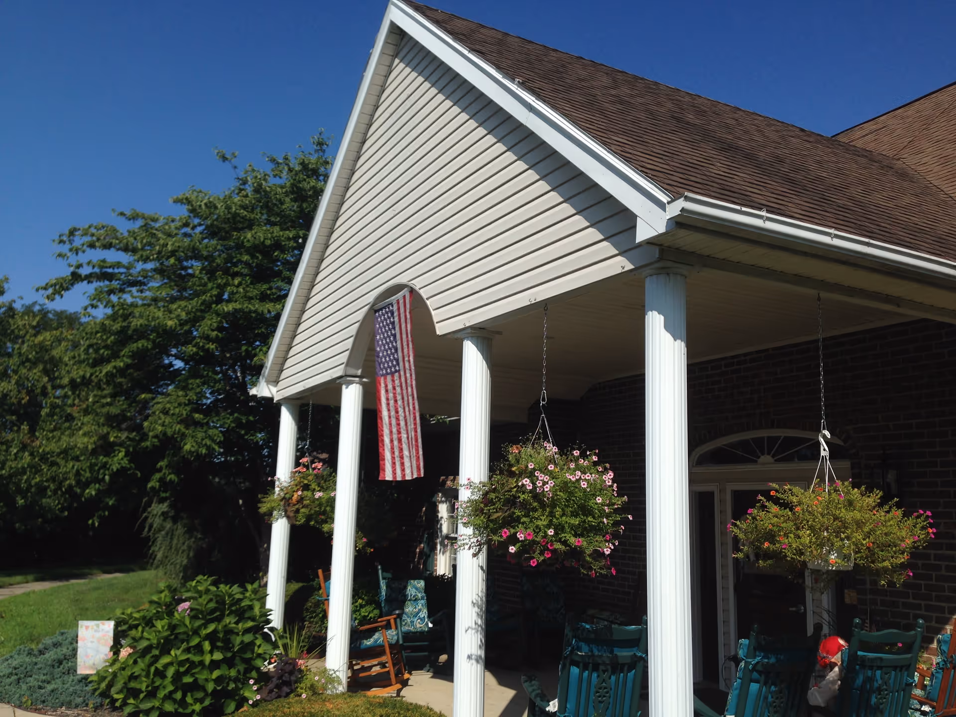 Front porch of a building with white columns and a peaked roof. Hanging flower baskets and an American flag are displayed. There are several rocking chairs on the porch and greenery surrounding the area under a clear blue sky.