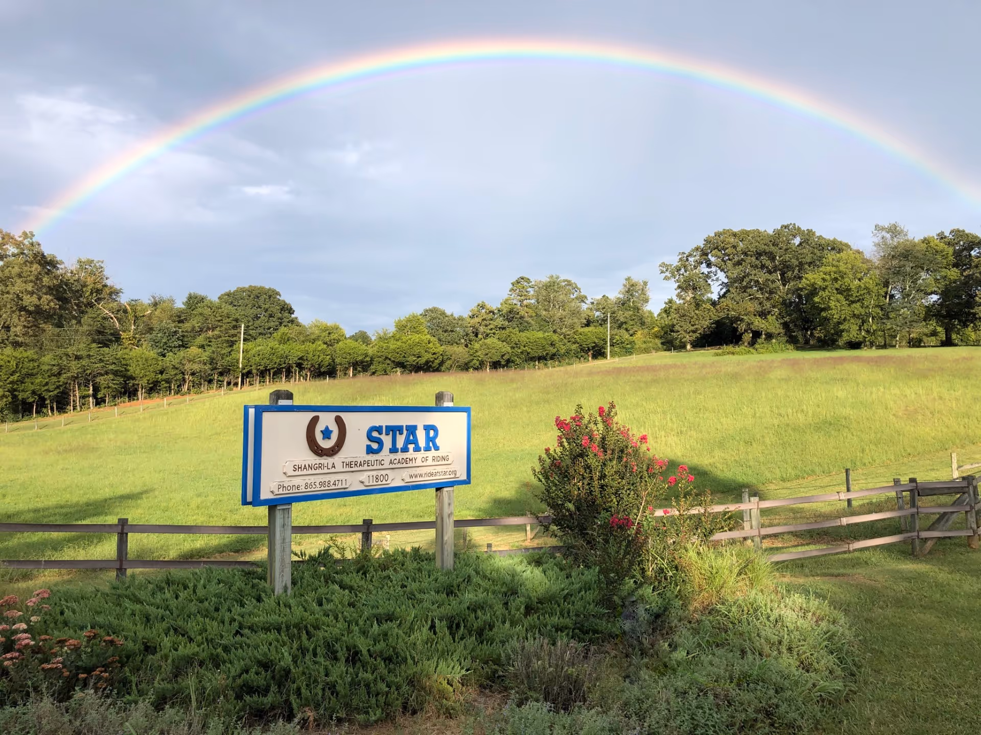 A rainbow arches over a grassy field with a wooden fence and a blue sign.
