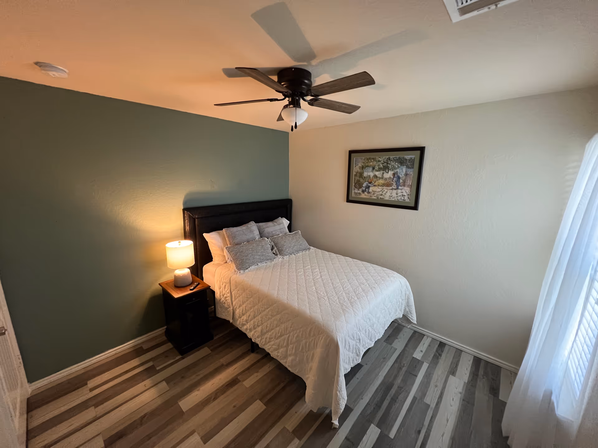 A cozy bedroom with a bed, nightstand, lamp, ceiling fan, and framed wall art.