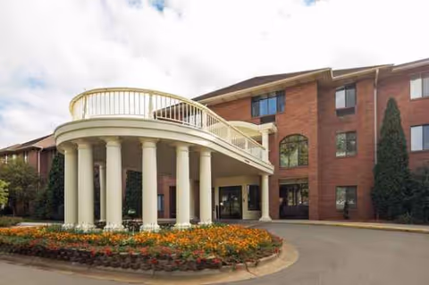 Brick senior living building with curved white portico and flower beds.