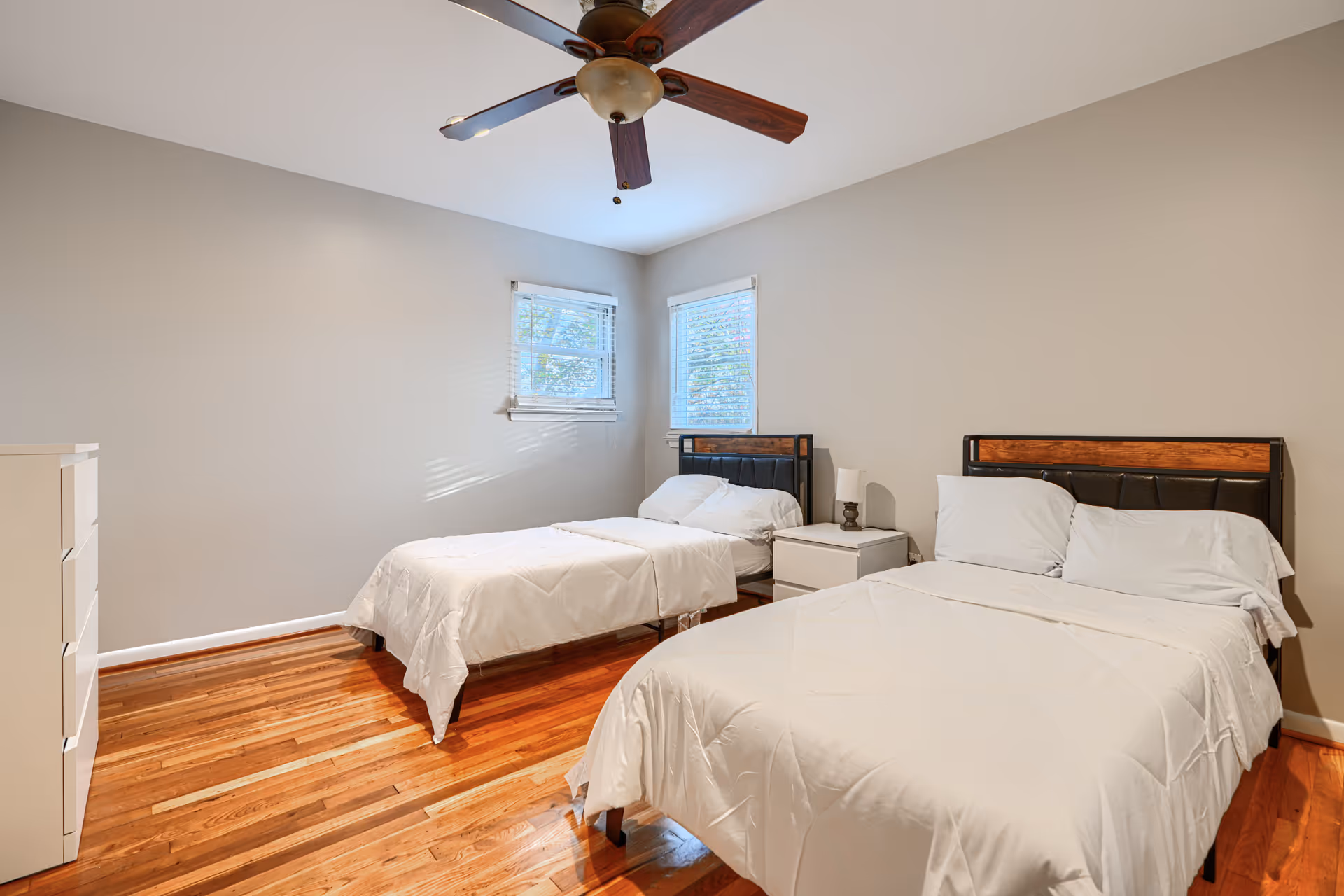 Two twin beds with white bedding, nightstand, dresser, and ceiling fan in bright bedroom.