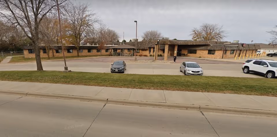 Front exterior of a long single-story brick building with a covered entrance, a small parking lot with cars, trees, sidewalk and a street in the foreground.