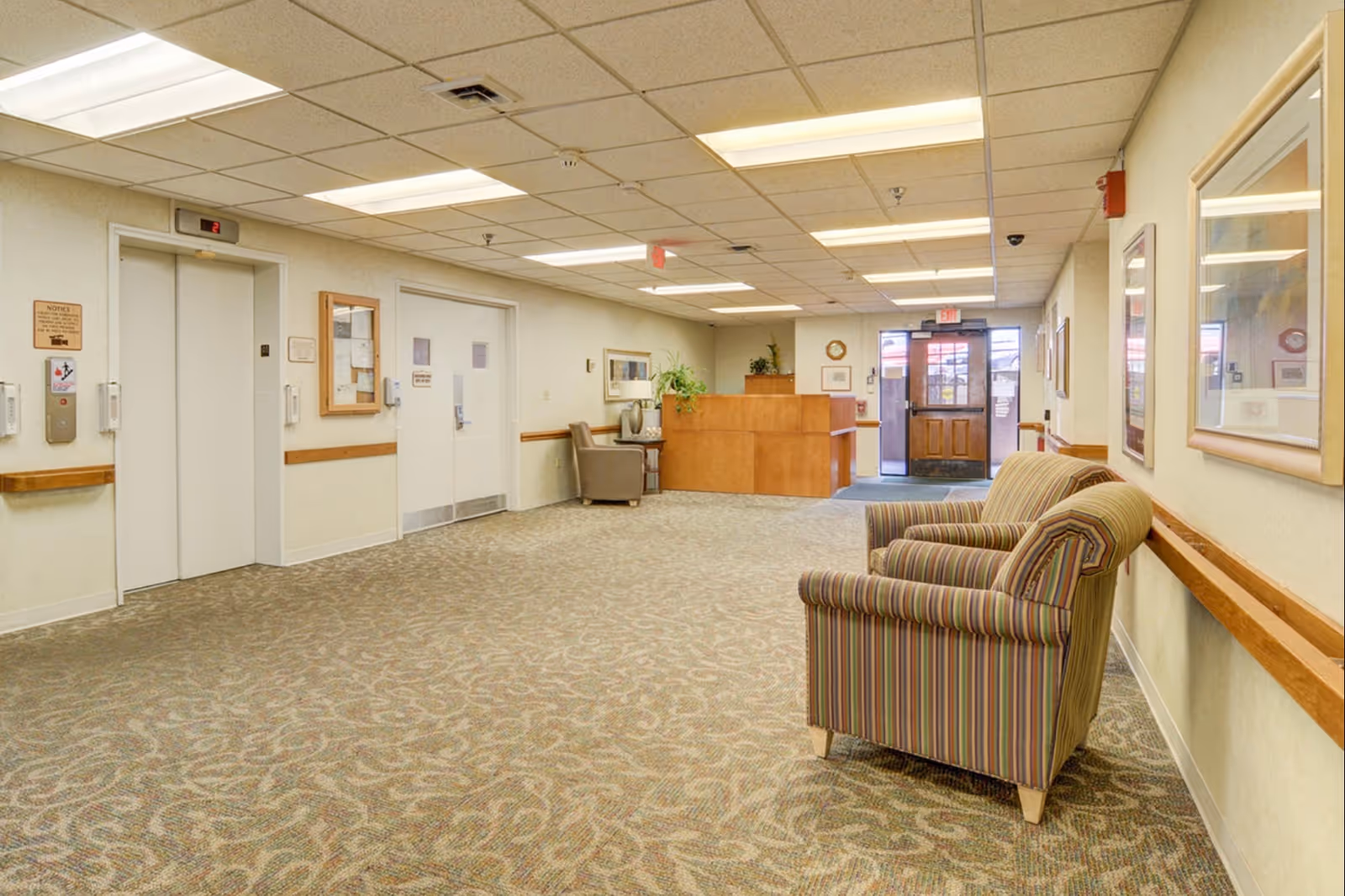 Lobby hallway with striped chairs, a reception desk, and doors in a senior care facility.