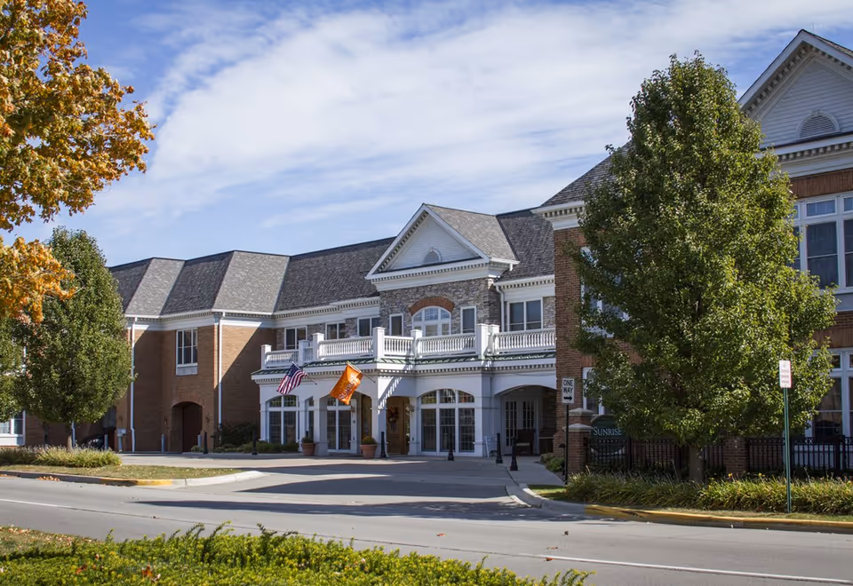 Sunrise of Grosse Pointe Woods senior living facility exterior with entrance, flags, and trees.