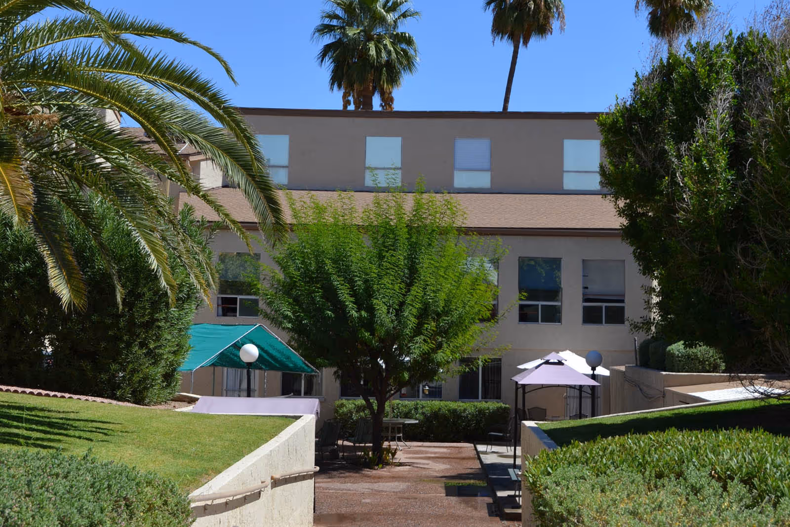 Courtyard with shaded seating, umbrellas, and trees in front of a multi-story senior living building.