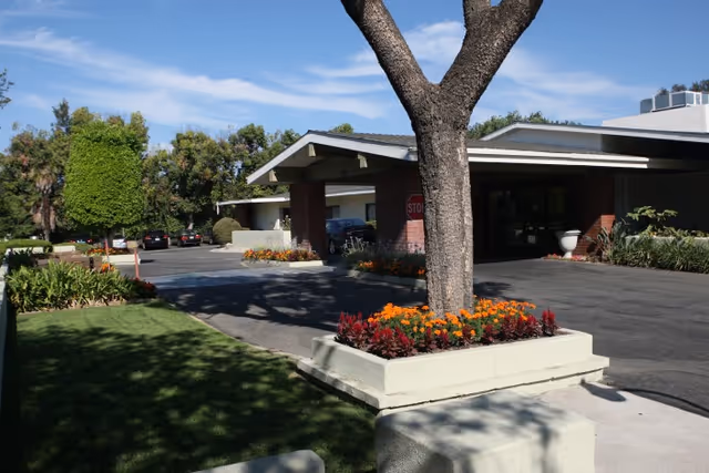 Driveway entrance of a care center with a covered porte-cochère, a tree in a raised planter, and landscaped flowerbeds.