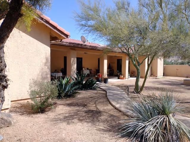 Outdoor view of a single-story building with a beige stucco exterior and a red tile roof. There is a curved concrete pathway leading to a covered patio area with several doors and potted plants. Desert landscaping with trees, shrubs, and dry soil surrounds the building under a clear blue sky.