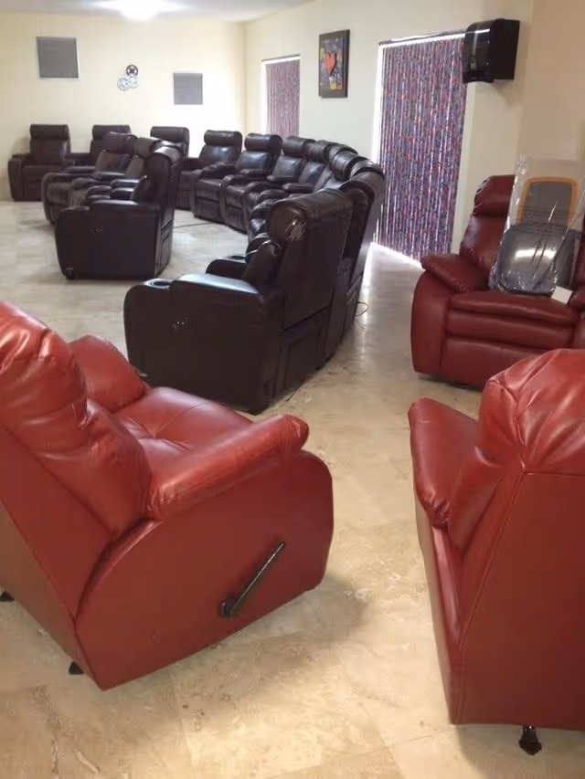 A room with multiple recliner chairs arranged in rows and a curved formation. The chairs are mostly dark brown with a few red ones. The room has light-colored tiled flooring, two windows with patterned curtains, a wall-mounted paper towel dispenser, and a framed artwork on the wall.