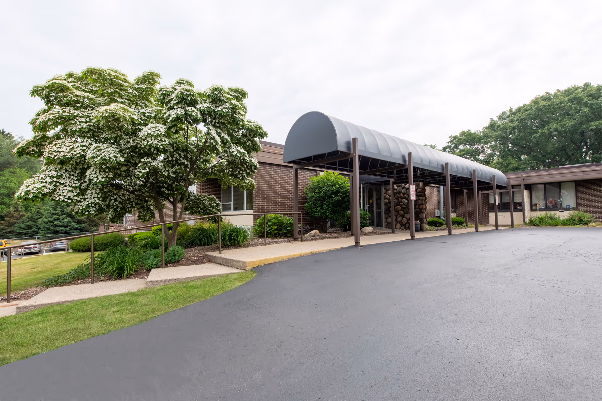 Exterior view of SKLD Leonard facility showing a single-story brick building with a covered entrance canopy. There is a paved driveway and a landscaped area with a flowering tree and green shrubs near the entrance.