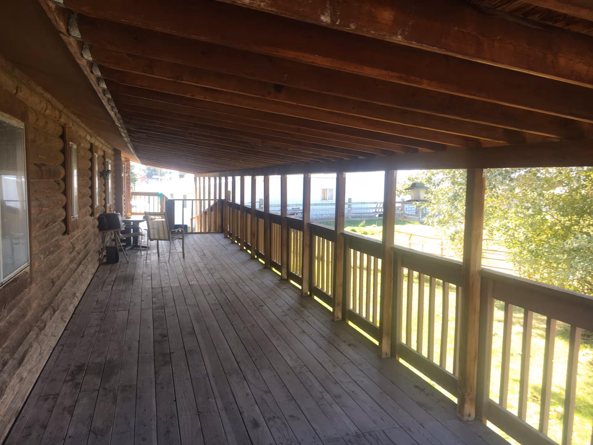 Long wooden covered outdoor deck with chairs along the railing beside a log-style facility.