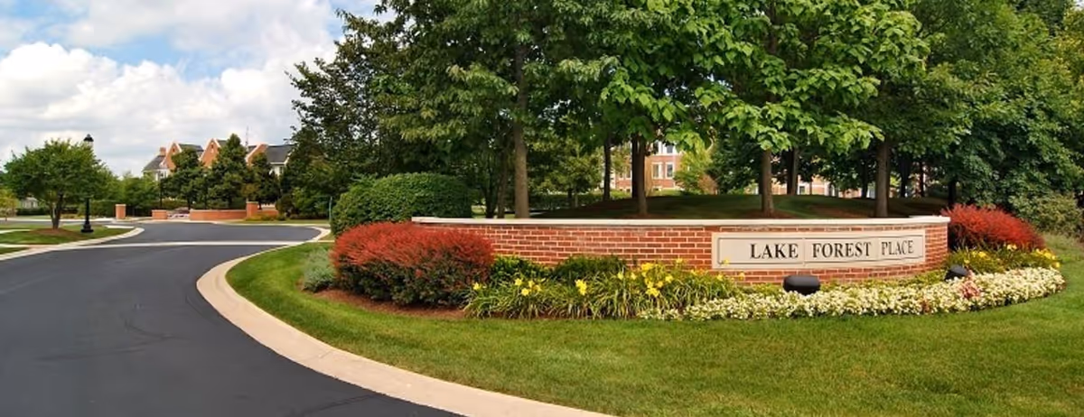 Curved brick entrance sign reading "LAKE FOREST PLACE" surrounded by landscaping, trees, and a paved driveway.