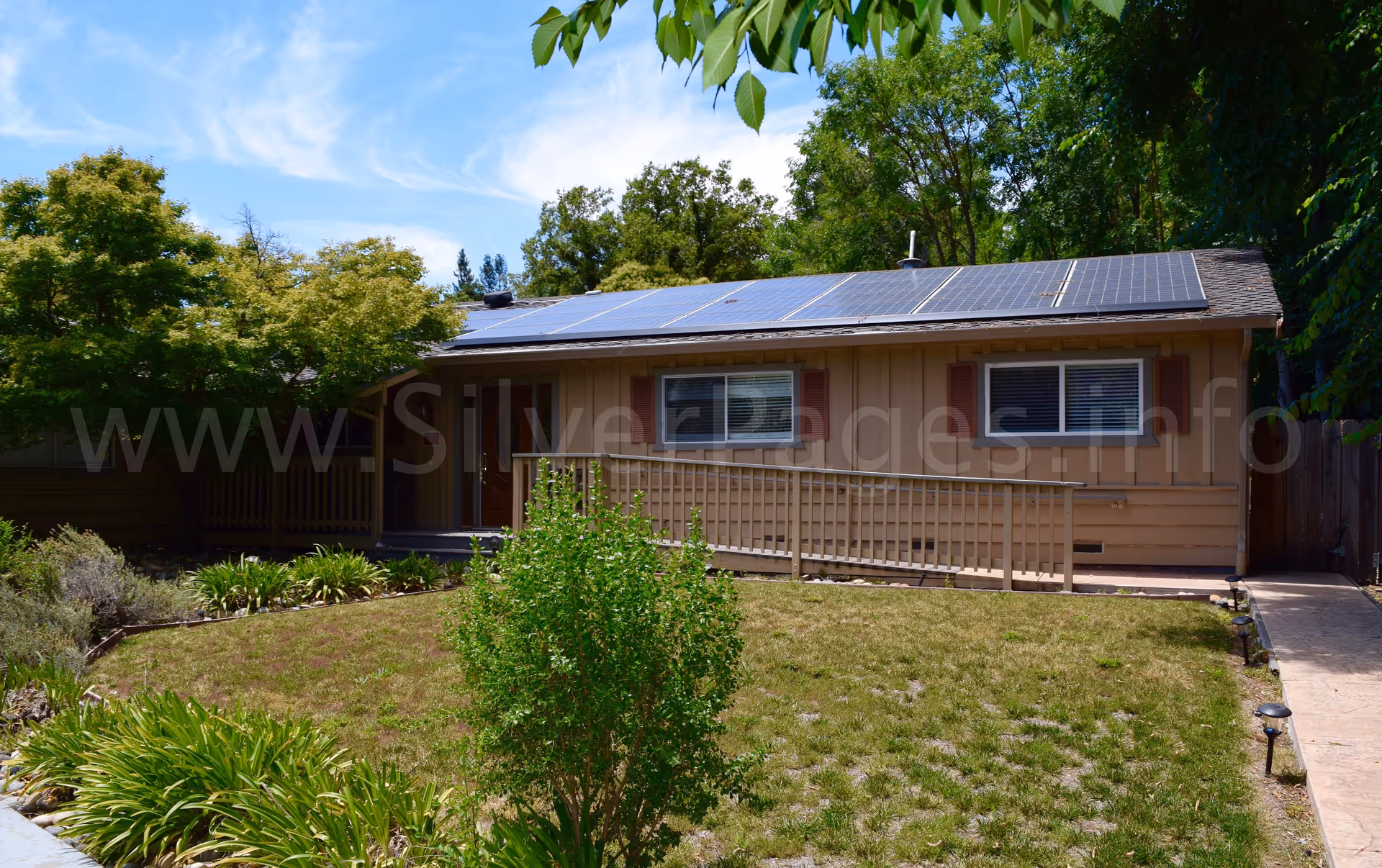 Single-story brown building with solar panels, a front porch railing, and lush green landscaping.