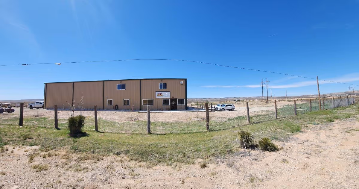 Desert landscape with a tan TLC Home Health Care building, fenced yard, and parked cars.