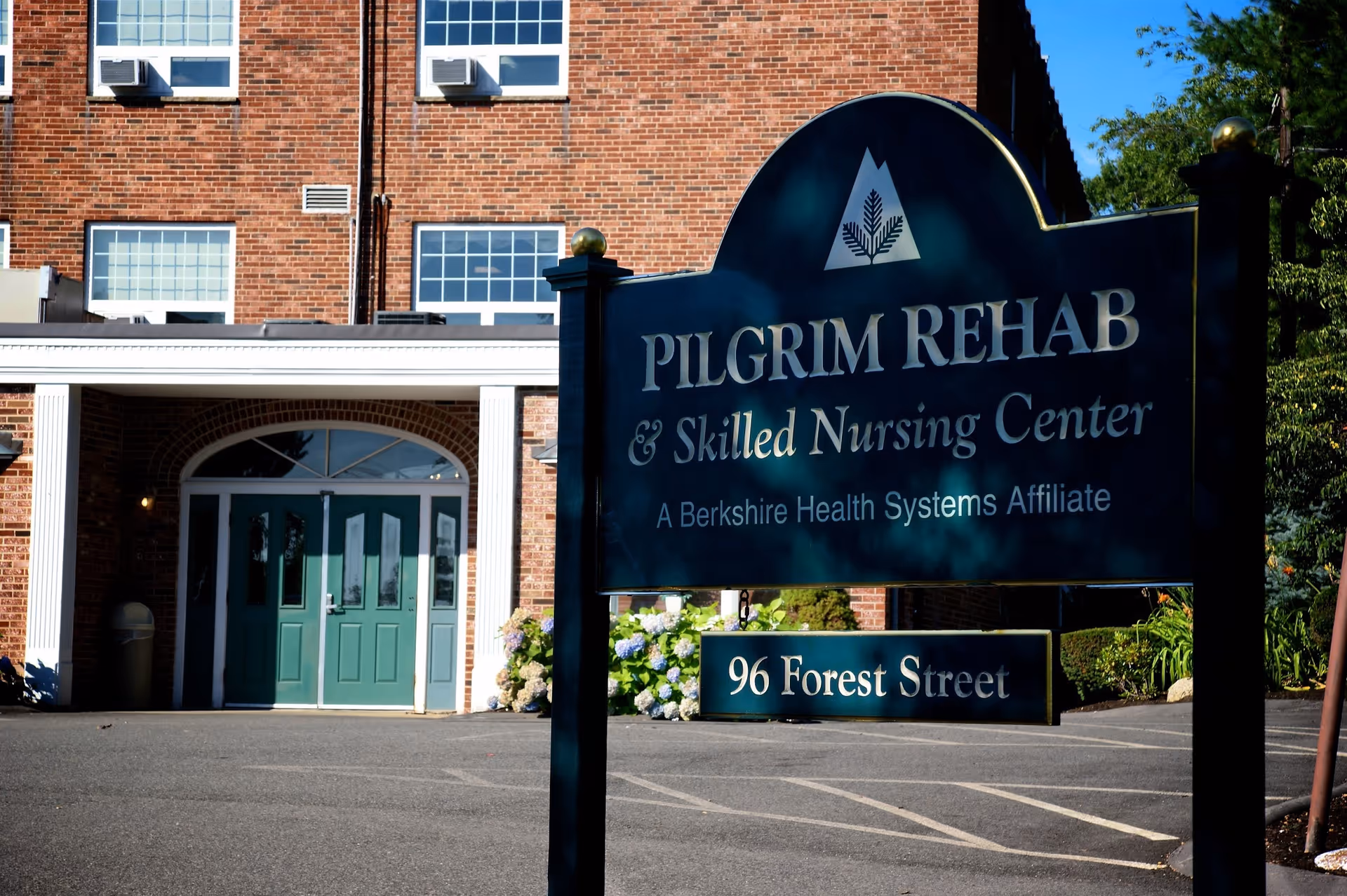 Exterior view of Pilgrim Rehab & Skilled Nursing Center building with a large sign in front displaying the facility name and address 96 Forest Street. The building is made of red brick with green double doors and white columns at the entrance.