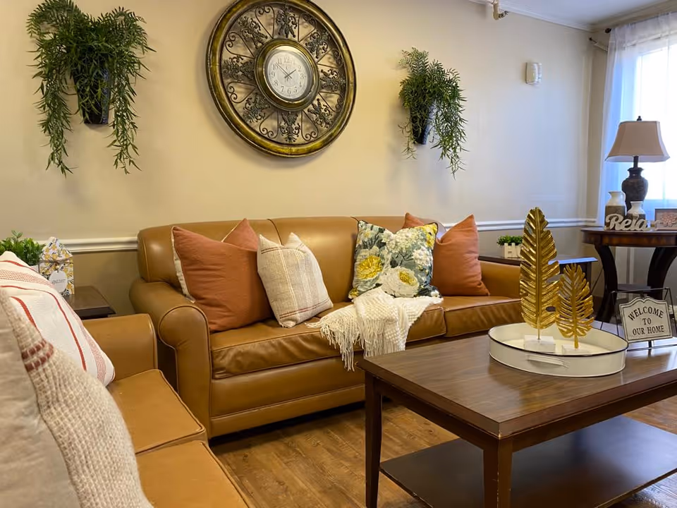 A cozy living room with a brown leather sofa and matching armchair adorned with various decorative pillows and a white throw blanket. A wooden coffee table in front holds a white tray with two gold leaf ornaments. On the wall above the sofa is a large decorative clock flanked by two hanging green plants. To the right, a side table with a lamp, small plants, and a 'Relax' sign is visible, along with a small sign that says 'Welcome to Our Home'. Light filters in through a window with sheer white curtains.