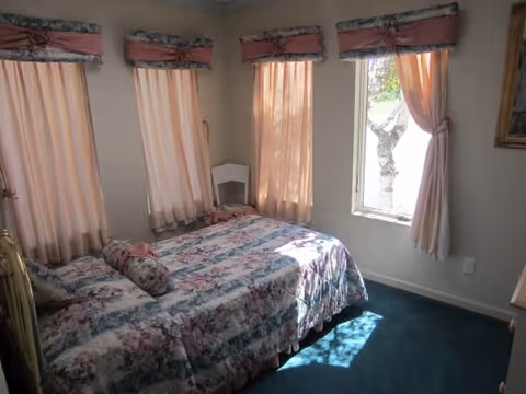 A small bedroom with floral bedspreads, pink curtains, and sunlight on blue carpet.