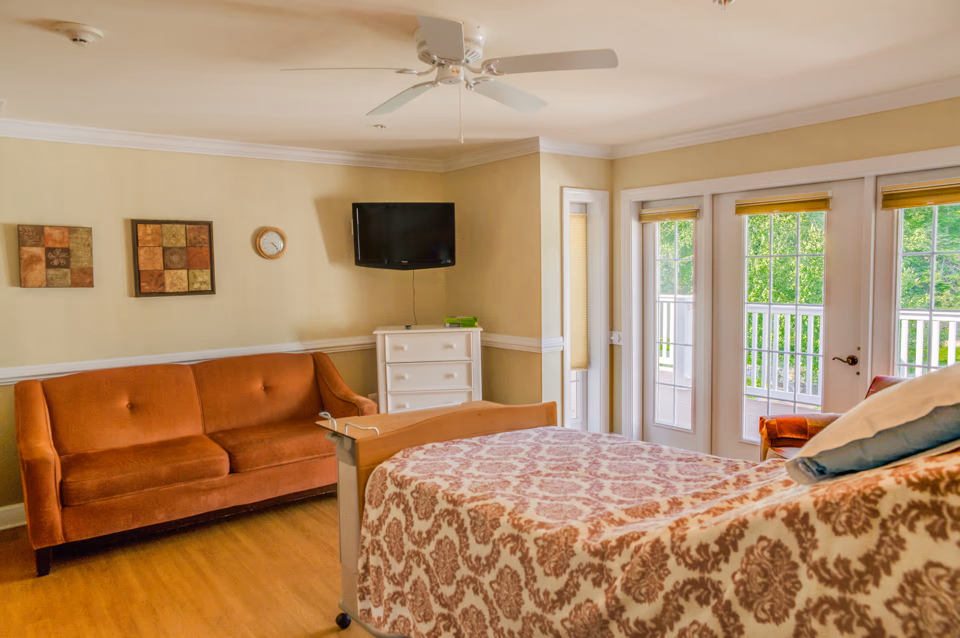 Spacious senior living bedroom with bed, orange sofa, dresser, mounted TV, and glass doors.