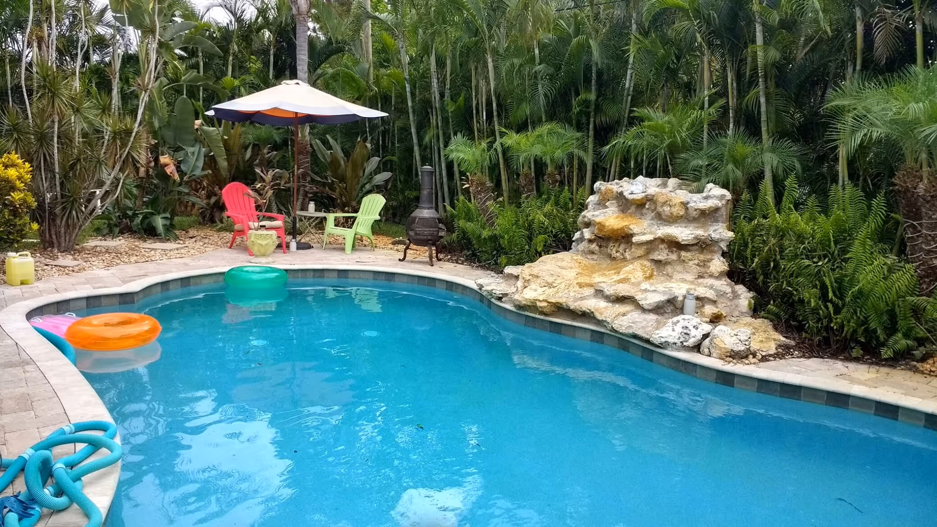 Outdoor pool area with lounge chairs, umbrella, rocks, and tropical plants.