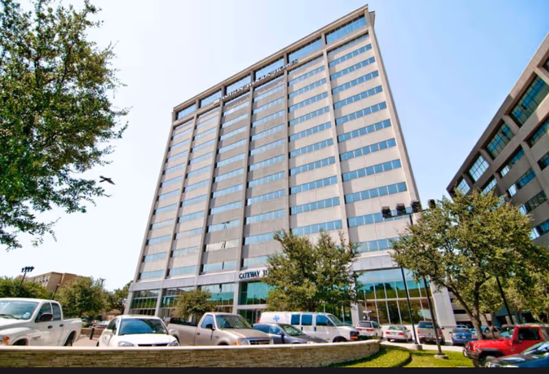 Tall, glass-fronted, multi-story building with a parking lot and trees in front.