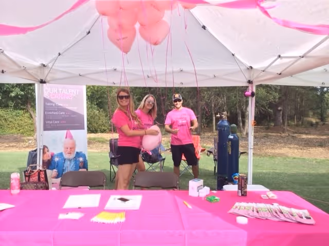 Three staff members in pink shirts stand under a canopy at a pink-decorated outdoor booth.