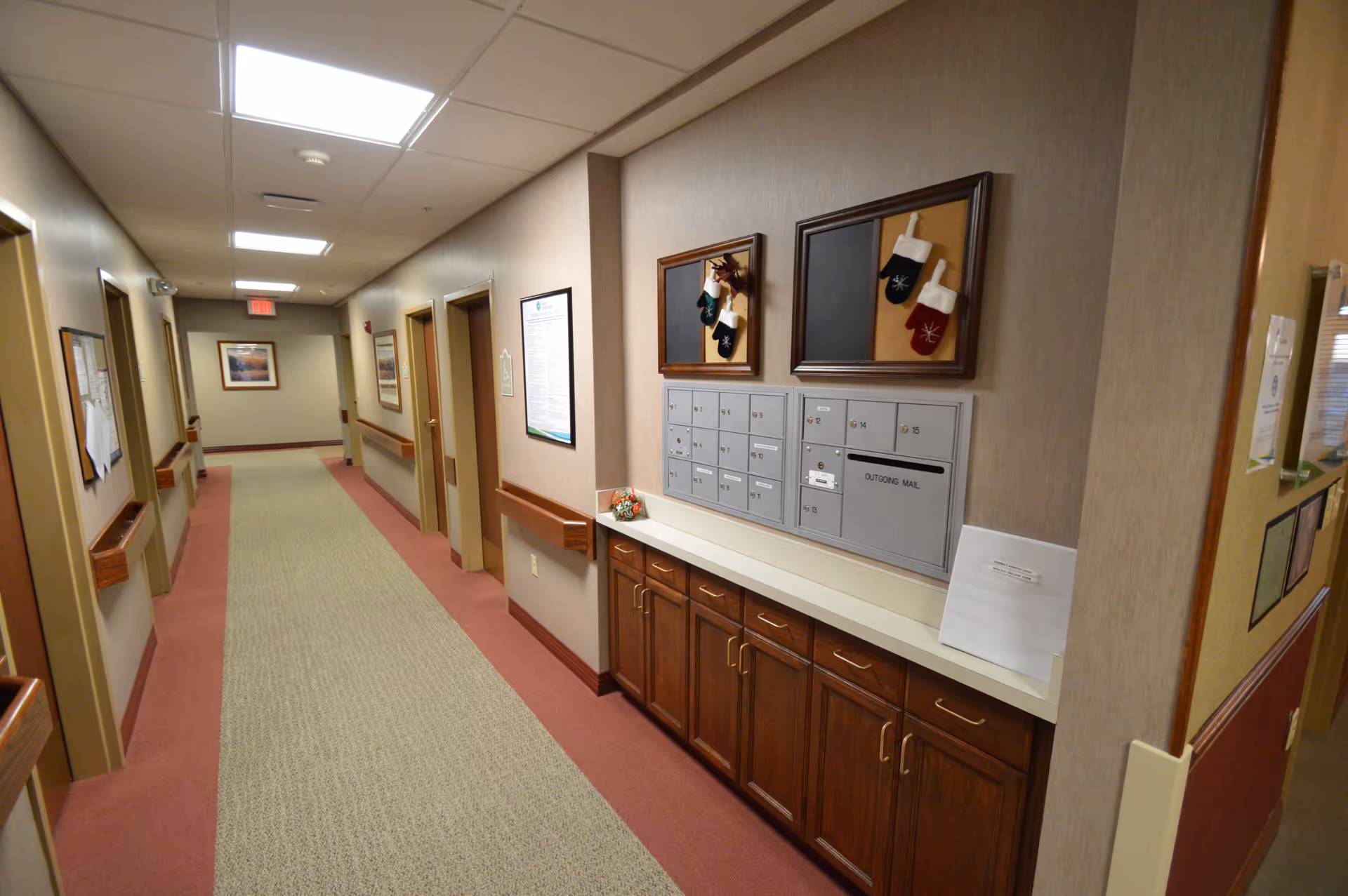 A well-lit hallway in a senior living facility with beige walls and carpeted floor. On the right side, there are multiple mailboxes mounted on the wall above a wooden cabinet. The hallway has several doors along the left and right walls, handrails, and framed pictures hanging on the walls. The ceiling has recessed lighting panels.