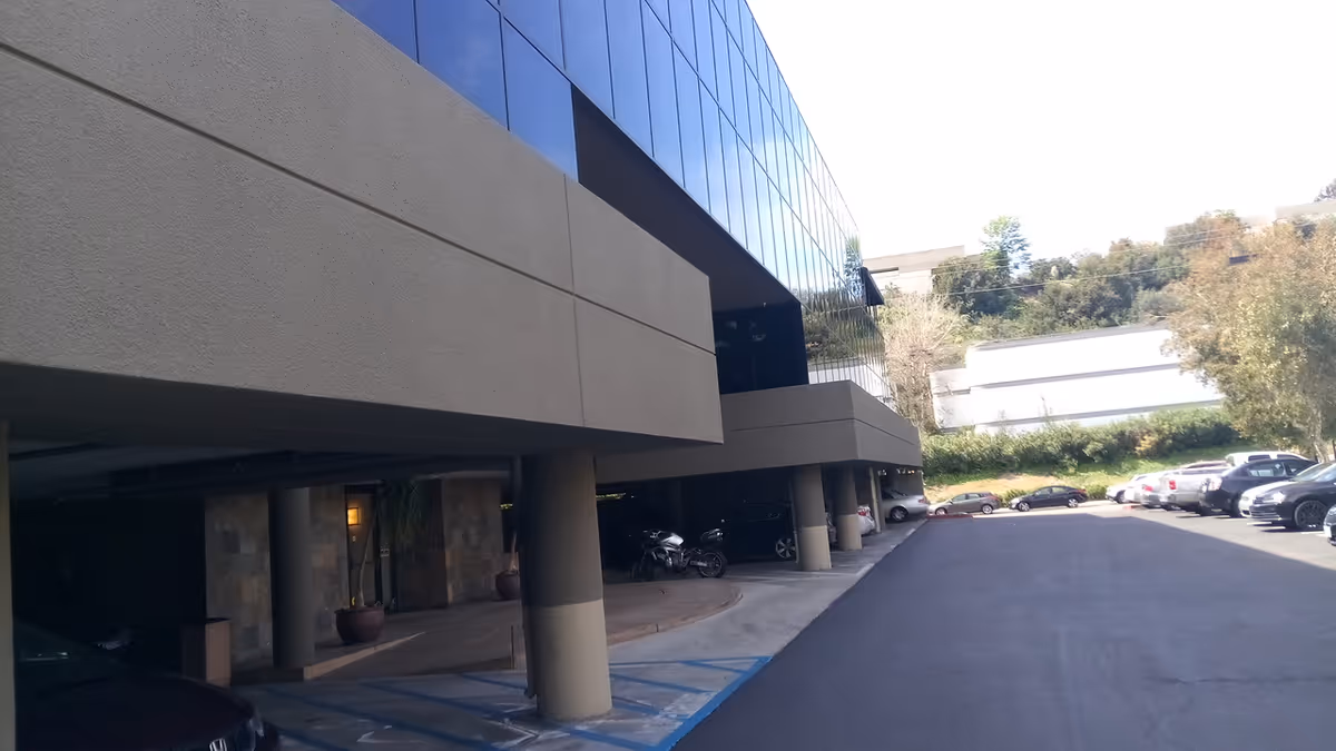 Modern building with glass facade, parking lot, and support columns along a driveway.