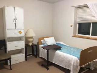 A small bedroom with a single bed covered with a white quilt and a blue blanket. There is a brown adjustable overbed table on wheels next to the bed. A white storage cabinet with drawers and a lamp on a small table are placed against the wall. A window with closed blinds and a sheer curtain is visible on the right side. A chair is partially visible on the left side of the image.