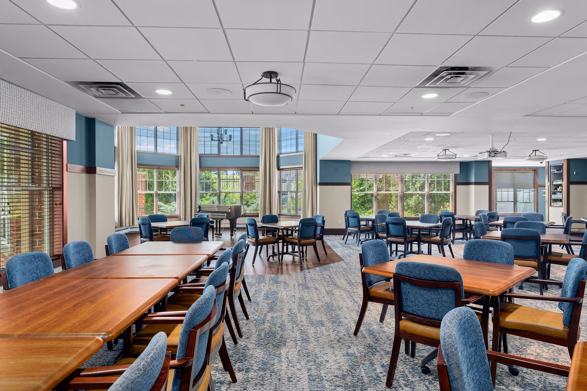 Spacious senior living dining area with multiple wooden tables and blue upholstered chairs.
