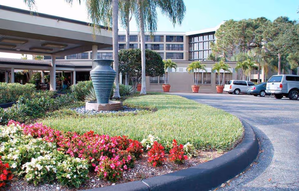 Front entrance of a senior living building with a circular landscaped driveway, a large decorative urn, palm trees, flower beds, and parked cars.