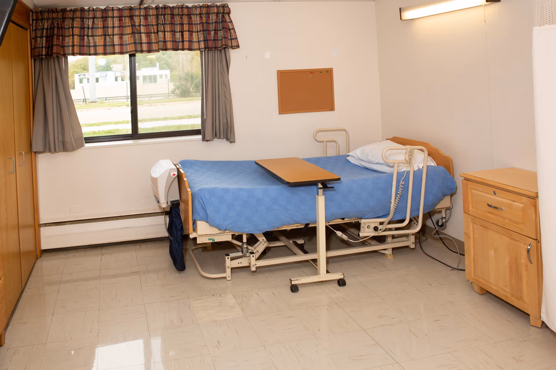 A simple bedroom in a senior living facility with a hospital-style bed covered with a blue blanket, a wooden bedside cabinet, a window with plaid valance and gray curtains, and a small cork bulletin board on the wall.