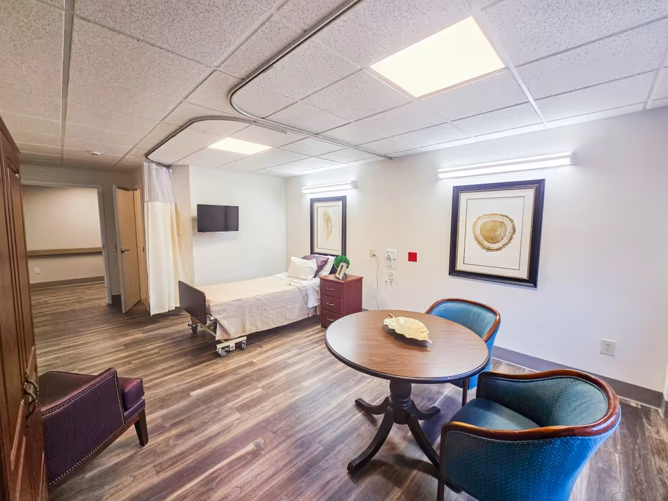 Single patient room with bed, privacy curtain, round table, chairs, and wall art.