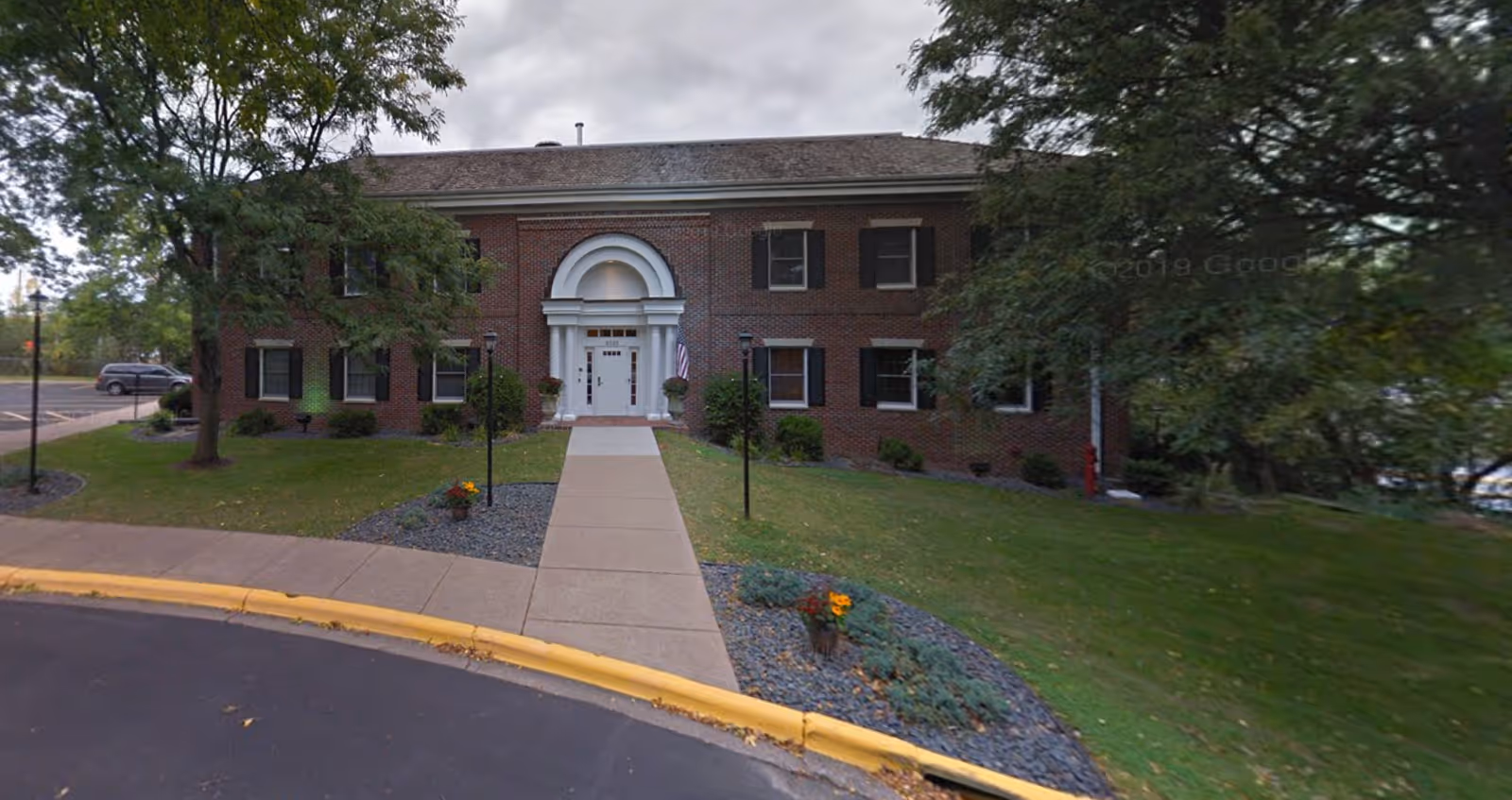Two-story brick building with a central arched entrance, walkway, lawn and trees in front.