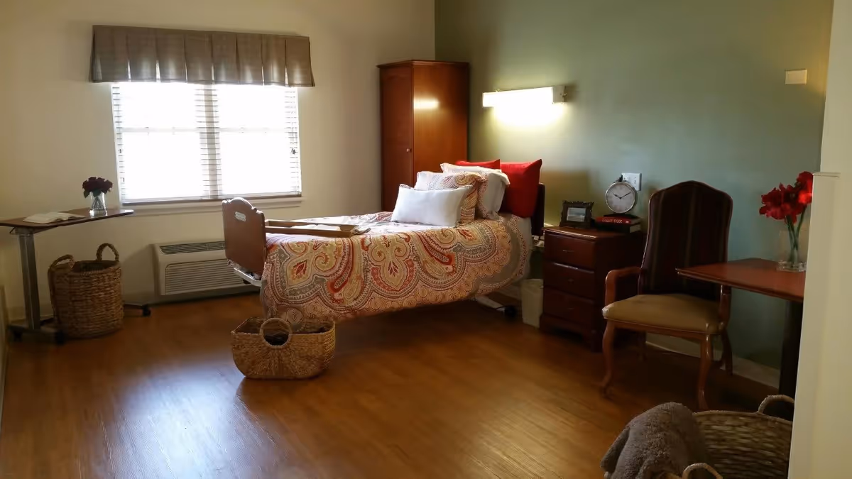Single furnished bedroom with a patterned bed, bedside dresser and chair, windows with blinds, and wooden flooring.