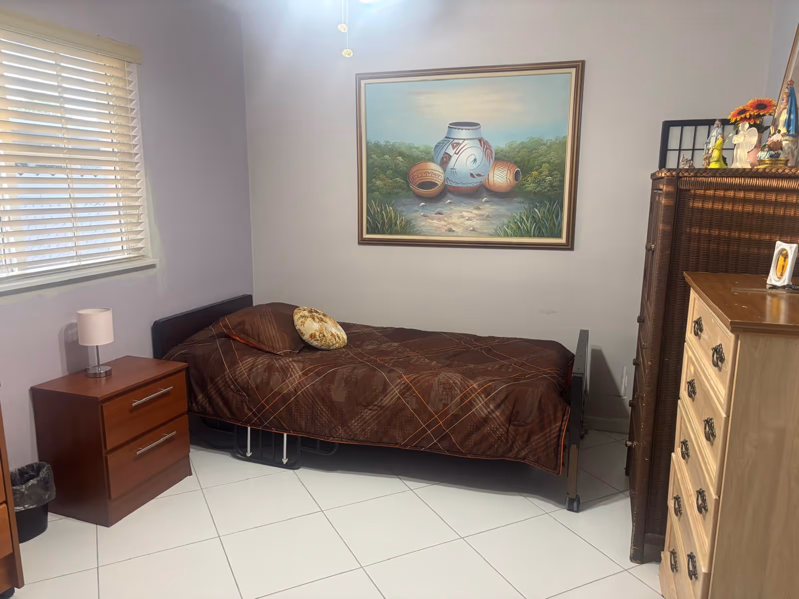 A bedroom in a senior facility featuring a bed, nightstand, dresser, and wall art.