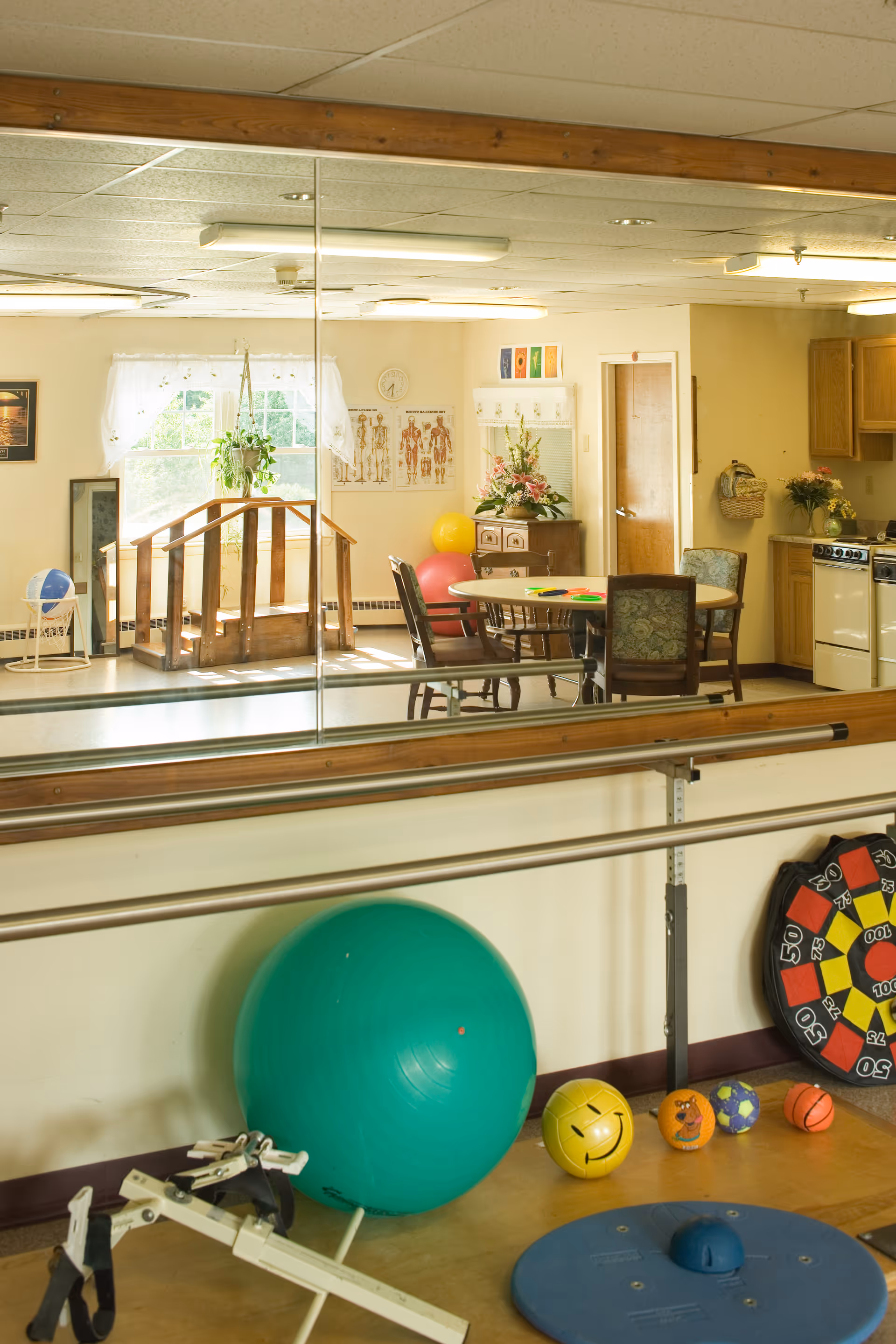 A therapy/activity room with parallel walking bars, a large green exercise ball, smaller therapy balls and a mirrored wall reflecting tables and chairs.
