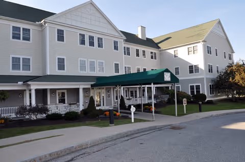 Exterior view of a large, multi-story senior living facility with white siding and a green roof canopy over the entrance. There is a paved driveway and landscaped bushes and trees around the building.