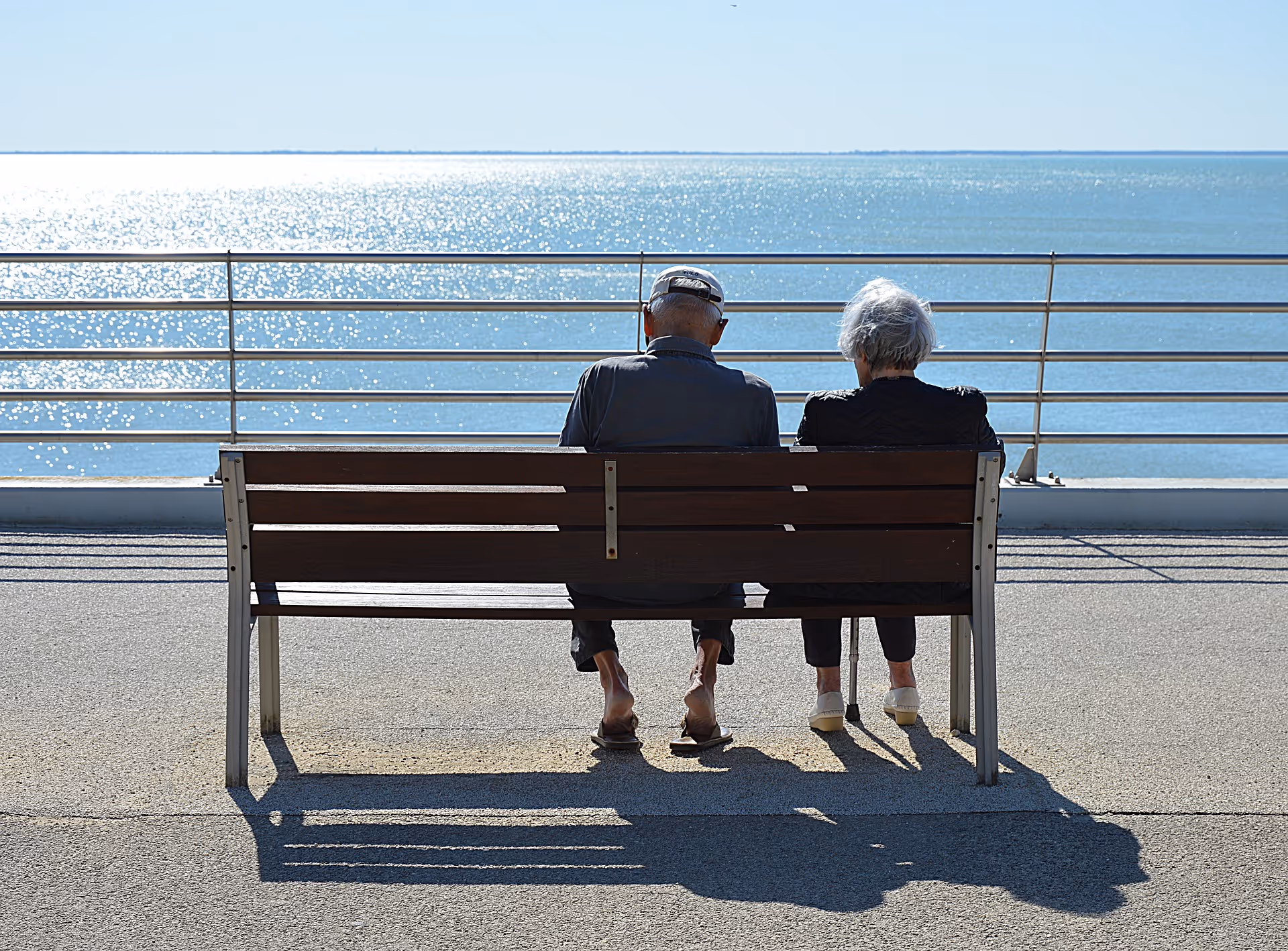 Two elderly people sit on a bench beside a railing, watching the ocean.
