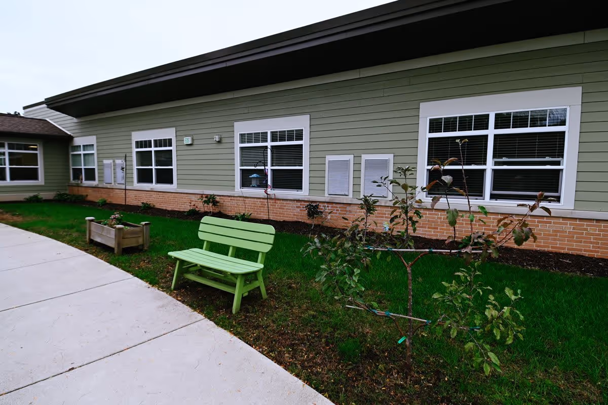 Green bench and planter beside a sidewalk in a small landscaped courtyard outside a single-story building with multiple windows.