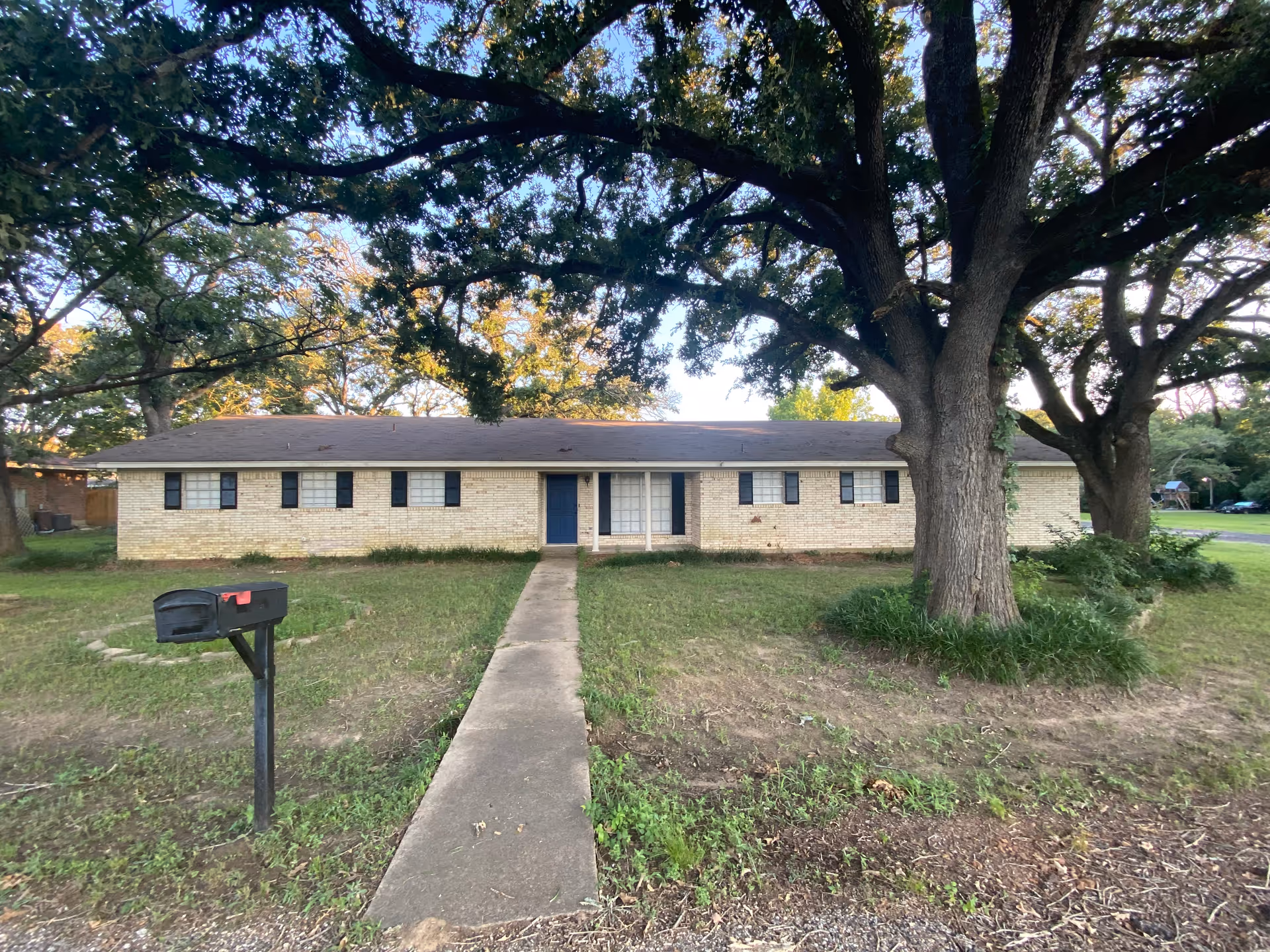 Single-story brick building with a blue front door and multiple windows with black shutters. A concrete walkway leads from the front door to a black mailbox on a post. Large trees with green foliage are in the front yard, providing shade.