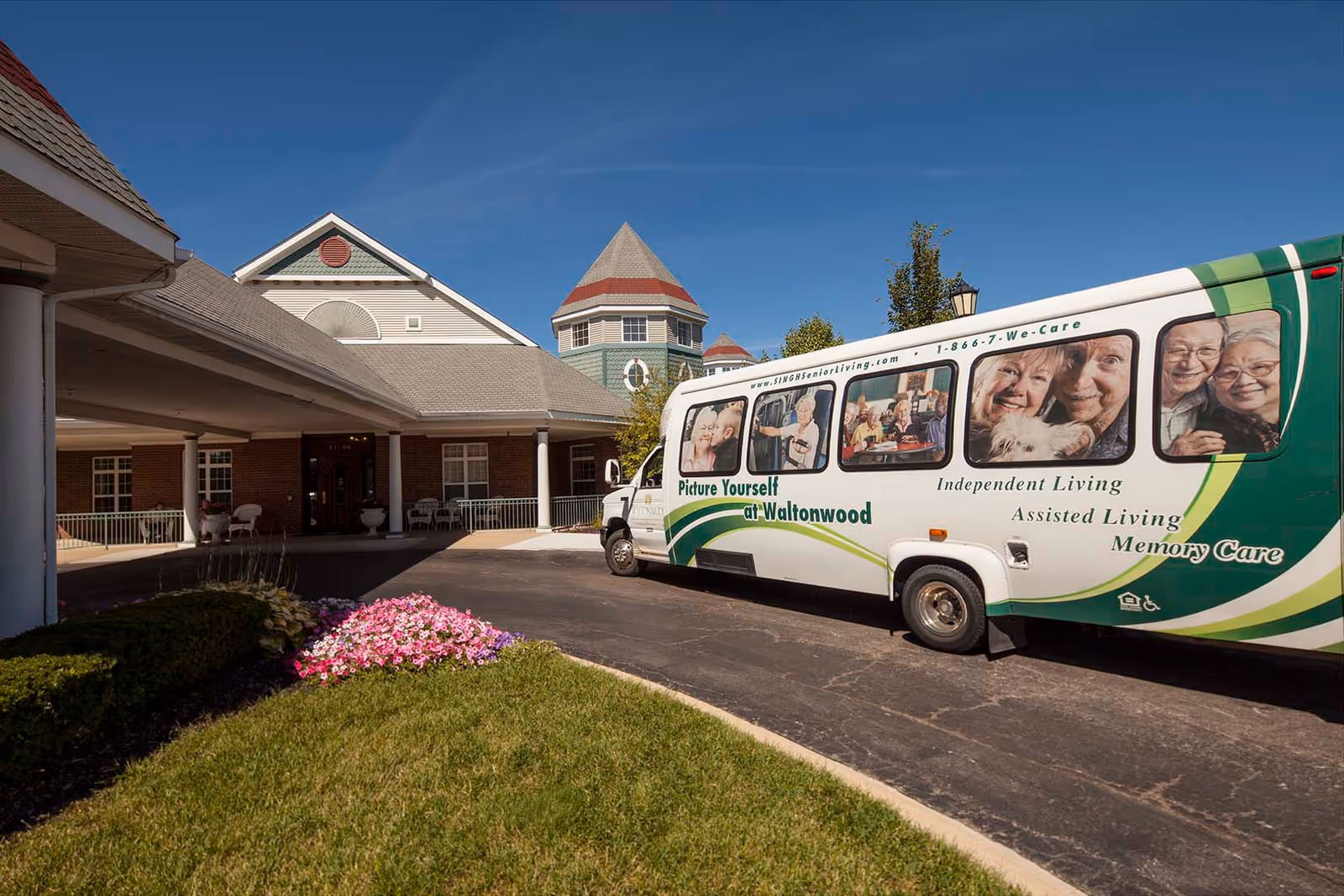 Exterior view of Waltonwood Cherry Hill senior living facility with a shuttle bus parked in front. The bus displays images of smiling elderly people and text promoting Independent Living, Assisted Living, and Memory Care services. The building has a covered entrance, brick and siding exterior, and a well-maintained lawn with flowers.