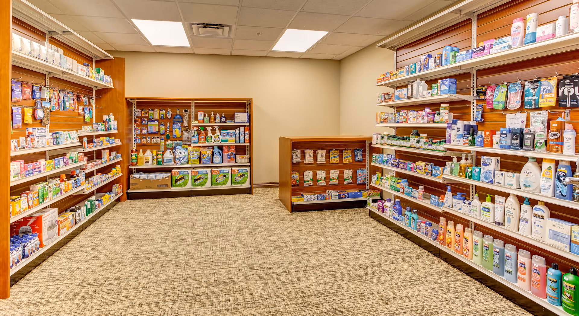 Interior view of a small retail shop area inside a facility, featuring shelves stocked with various personal care products, household cleaning supplies, and over-the-counter medications. The room has beige walls, a carpeted floor, and fluorescent ceiling lights.