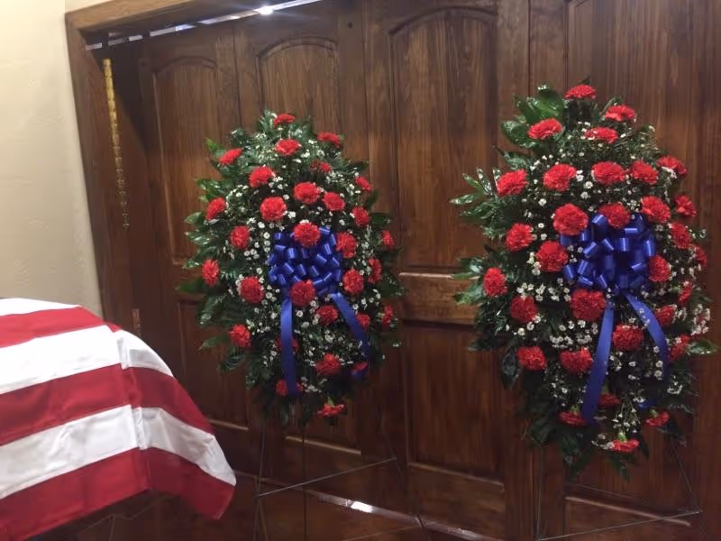 Two tall funeral wreaths with red roses and blue ribbons stand by wooden doors inside.